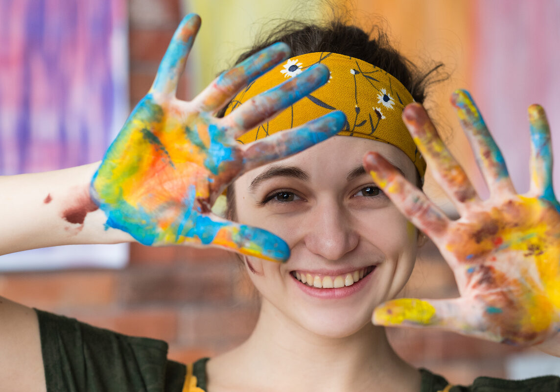 Art therapy. Closeup portrait of young female artist having fun in studio, smiling, showing hands dirty with paint.